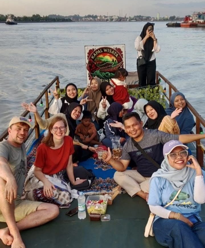 Sunset river cruise with traditional Indonesian wooden boat silhouetted against golden orange sky reflecting on water with palm trees along the riverbank
